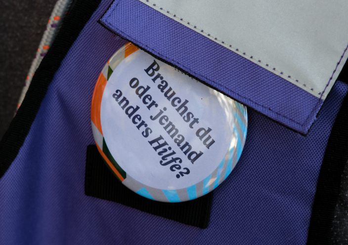 A round badge with the inscription 'Do you or someone else need help?' attached to a purple bag. The button is multicolored with blue and orange stripes on the edge.