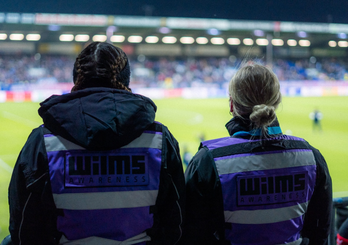 Two people seen from behind sitting on a grandstand, watching a football game. They wear jackets with the inscription 'Wilms Awareness' in purple and black. The illuminated football field with blurred spectators is visible in the background.