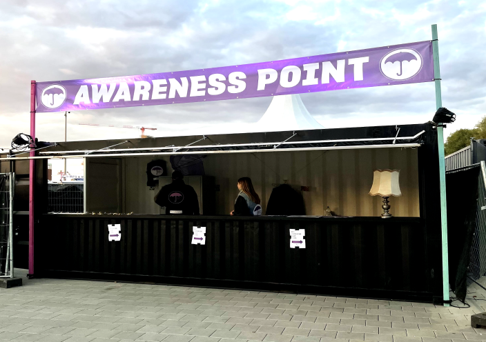 An outdoor information stand with the inscription 'AWARENESS POINT' on a purple banner. The stand has a black counter, behind which two people are standing. On the counter, there is a laptop and a desk lamp. The stand is illuminated, and the sky in the background shows twilight.