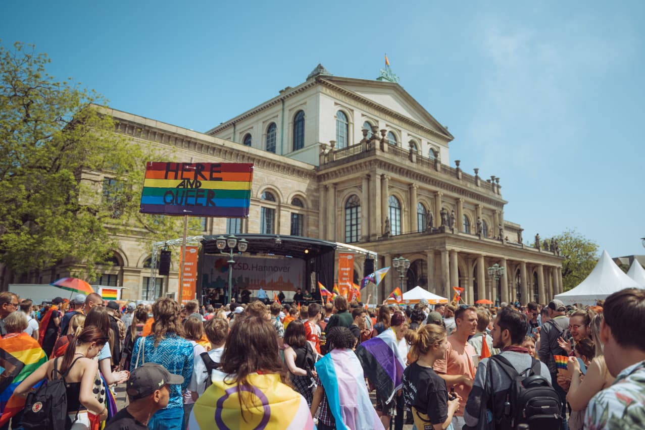 Pride march in front of Hanover's opera house with a rainbow “Here and queer” sign and crowds carrying flags.