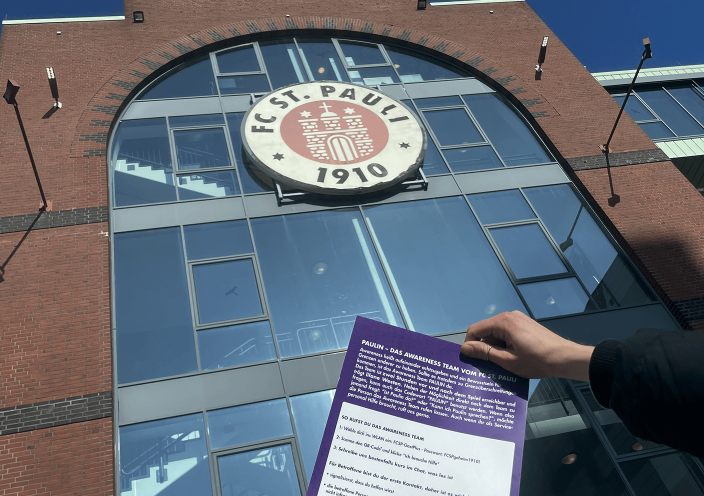 A hand holding a purple info sheet in front of the FC St. Pauli stadium entrance.