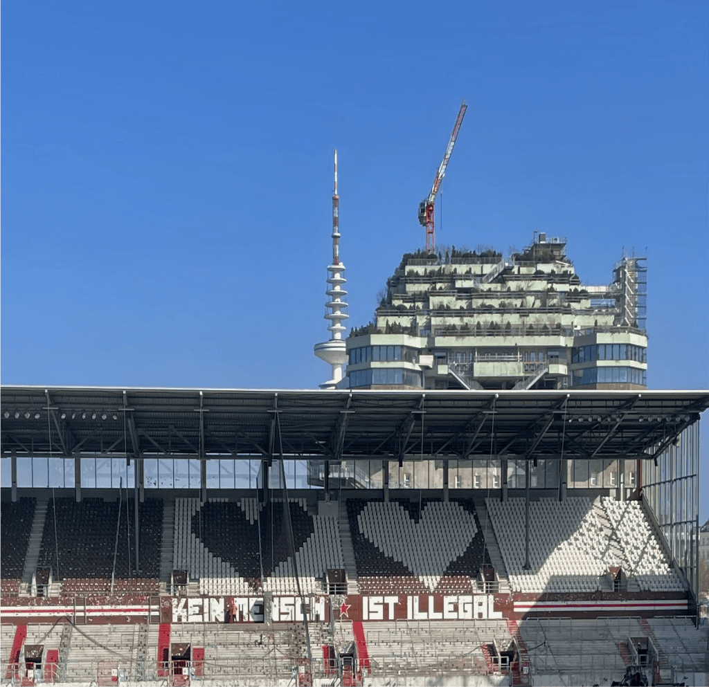 Stand at Millerntor stadium with the slogan “No human is illegal” and a city skyline backdrop.