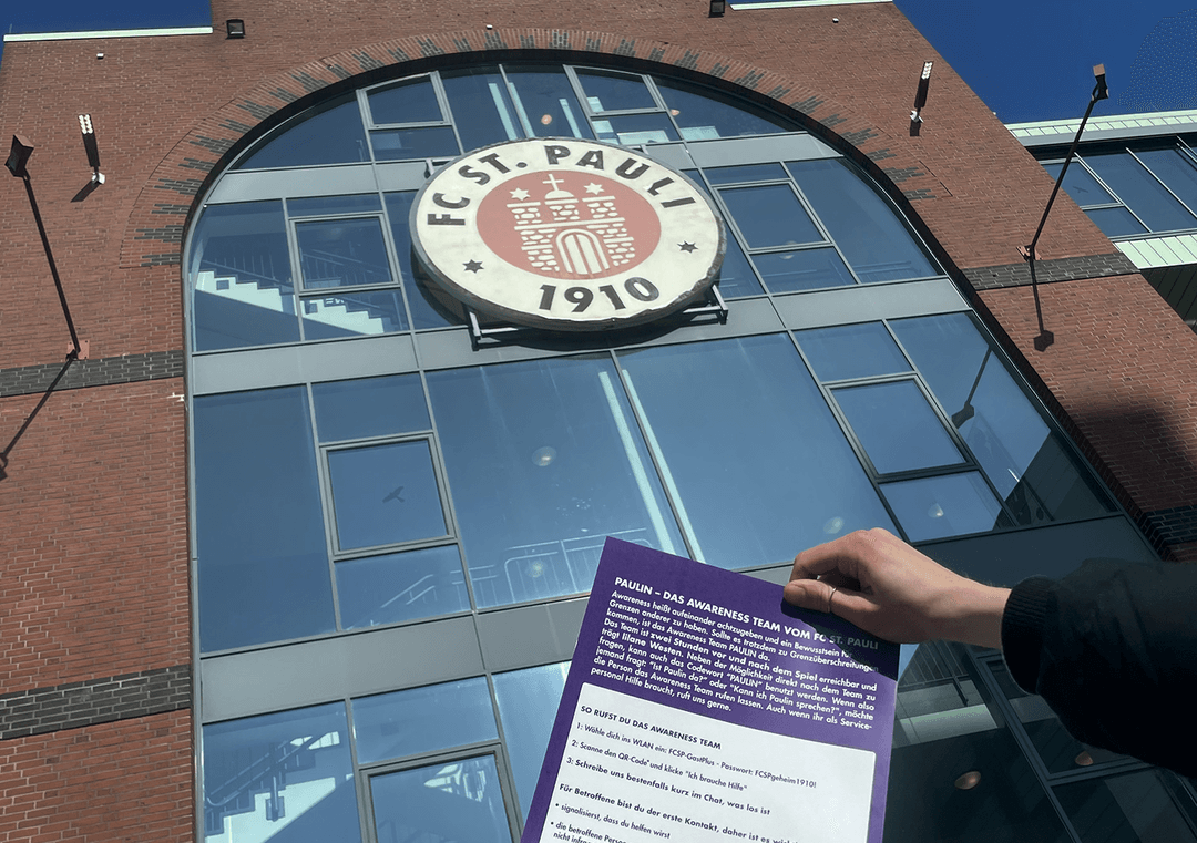 Hand holding a lavender information leaflet in front of the entrance of the FC St. Pauli stadium with large club logo and the inscription „1910“.