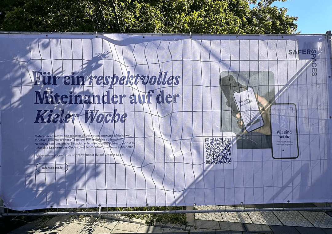 Banner with text: „Für ein respektvolles Miteinander auf der Kieler Woche“, QR-Code und Handy mit App „Wir sind bei dir!“.