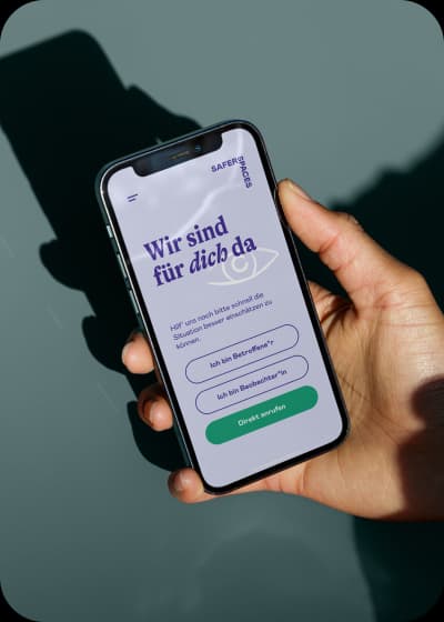 Eine Hand hält ein Smartphone. Auf dem Display ist die saferspaces WebApp geöffnet. Oben steht 'Wir sind für dich da'. Da drunter sind drei Auswahlmöglichkeiten: 1. Ich bin Betroffene*r, 2. Ich bin Boebachter*in, 3. Direkt anrufen.