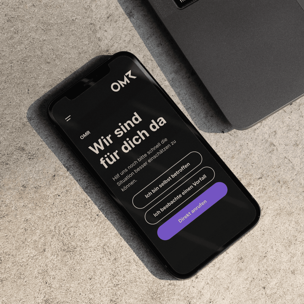 Smartphone on a stone surface showing the Saferspaces reporting interface branded for OMR. The heading reads 'Wir sind für dich da' with three action buttons.